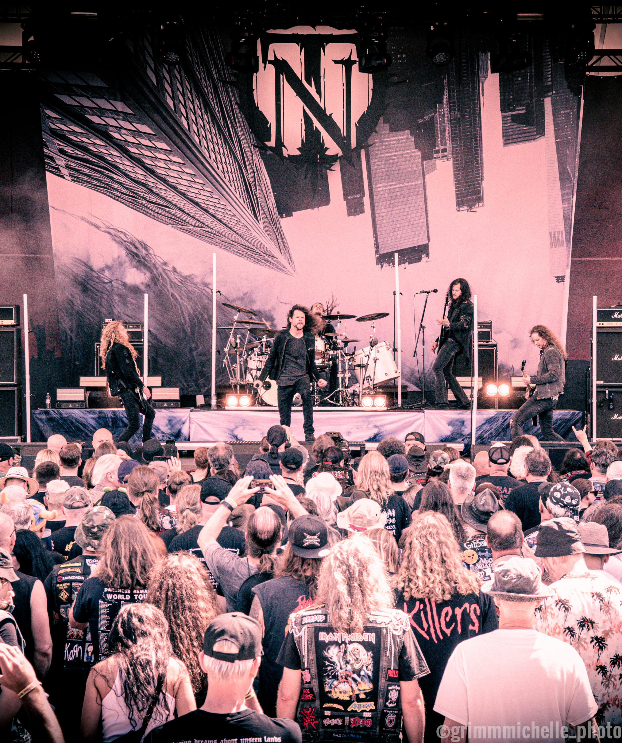 A live concert photo with a pinkish tint showing a heavy metal band performing on an outdoor stage. In the foreground, a large crowd of fans with their backs to the camera watches the performance. On stage, five band members are playing their instruments under a large backdrop featuring a stylized logo and skyscraper imagery.